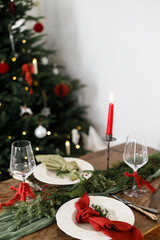 Merry Christmas! Stylish table setting for christmas family dinner. Modern plate and cutlery with napkin, glassware, green and red ribbons, fir branches and candle on rustic table at xmas tree