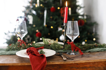 Stylish christmas table setting with modern plate, red and green napkins, cutlery, glass, fir branches, berries, candle on background christmas tree. Merry Christmas and Happy Holidays!