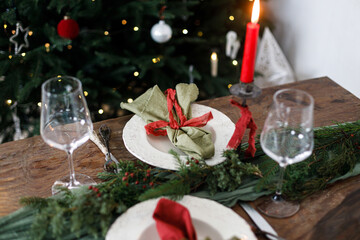 Stylish christmas table setting for family dinner. Elegant plate with napkins, cutlery, glassware, ribbons, green fir branches, red berries and candle against xmas tree lights. Holiday time