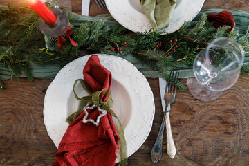 Merry Christmas! Stylish table setting for christmas family dinner. Modern plate and cutlery with napkin, glassware, green and red ribbons, fir branches and candle on rustic table flat lay