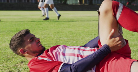 Obraz premium Wincing male soccer player clutching thigh on grass pitch during match, with red-and-white kit