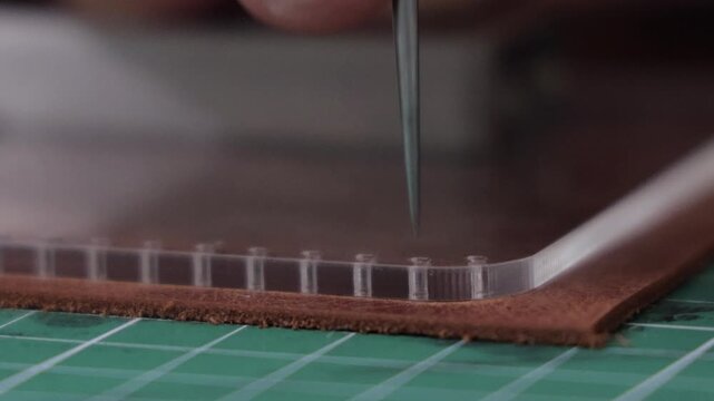 the meticulous process of leather crafting, featuring a hand using an awl to punch holes in a layered brown leather piece over a transparent plastic sheet, set on a green cutting mat