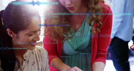 Leaning women collaborating over tablet at bright office desk, pointing at screen and documents