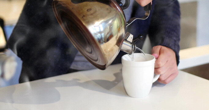 Pouring man wearing dark jacket tilting stainless steel kettle into ceramic mug at cafe counter