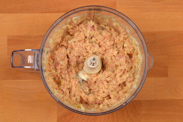 An overhead view of a bowl with a mixture of ground chicken meat and spices, prepared for making homemade hot dogs, inside a food processor on a wooden surface