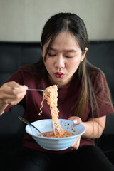 Asian woman sitting at home in sofa eating instant noodles from bowl