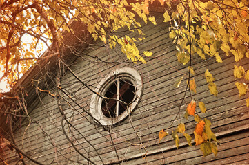 Old wooden house with a round attic window framed by autumn trees with golden leaves, vintage tones...