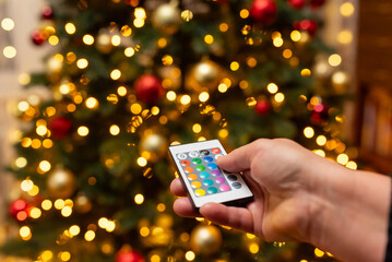 A person turning on Christmas lights with a remote control. A hand is holding a small remote control and pressing a button to turn on the lights on a Christmas tree