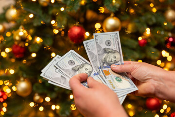 A person counting dollars against the background of a Christmas tree. Woman counting money near the Christmas tree, idea of big spending on gifts, decorations for New Year holidays