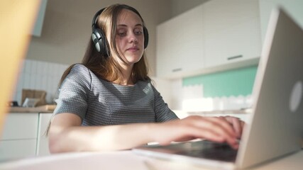 Working on laptop with headphone reaching hand toward viewer while woman types on laptop keyboard at kitchen desk wearing watch and headset showing online work and gesture during remote work session