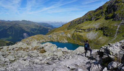 Adventurous hiker standing on rocky terrain overlooking a serene mountain lake, surrounded by lush greenery and majestic peaks under a clear blue sky, capturing the essence of exploration