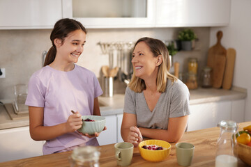 A mother and daughter are smiling and enjoying each other's company in a bright kitchen. The daughter holds a bowl of fruit, while the mother attentively listens and laughs.