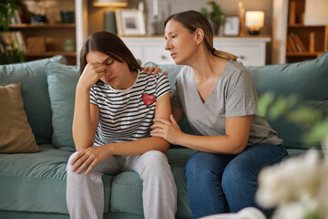 A mother comforts her teenage daughter sitting on a sofa in a warm living room, discussing personal struggles and emotions in an intimate, supportive atmosphere.