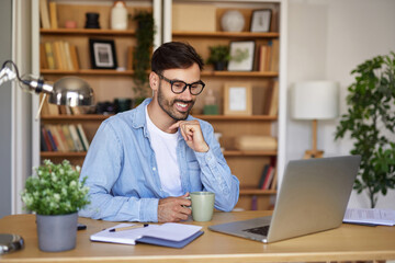 A young man is focused on his laptop in a well-organized home office, sipping coffee. The atmosphere is bright and inviting with plants and books around him.