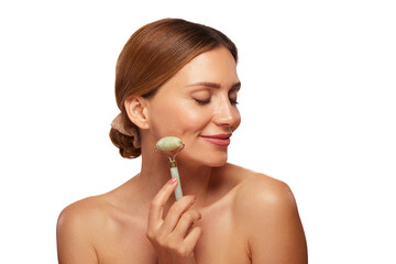 A woman smiles while gently using a jade roller on her cheek as part of her skin care routine. She...