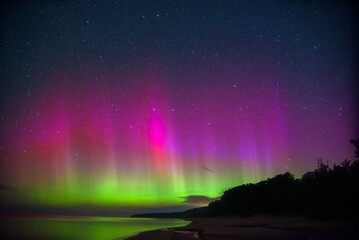 Bright pink and green aurora borealis lights dance across a starry night sky, illuminating the horizon over a calm coastal beach