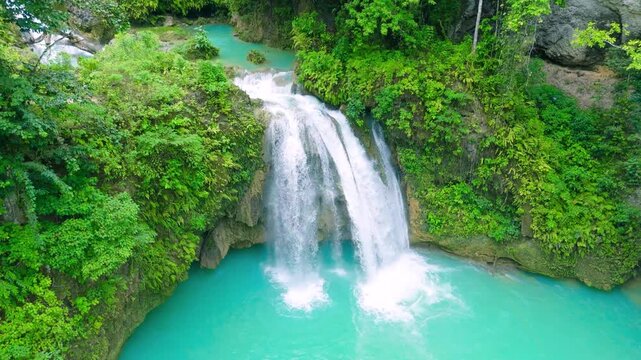 Aerial view of a multi-tiered waterfall cascading into a turquoise pool, with lush green jungle foliage and a person swimming, Kawasan Falls, Cebu.
