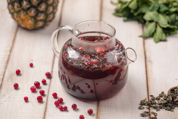 Glass teapot with hot berry tea and cranberries on wooden surface, next to mint and dried tea leaves