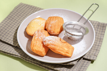 Plate with delicious madeleines, sieve and powdered sugar on green background