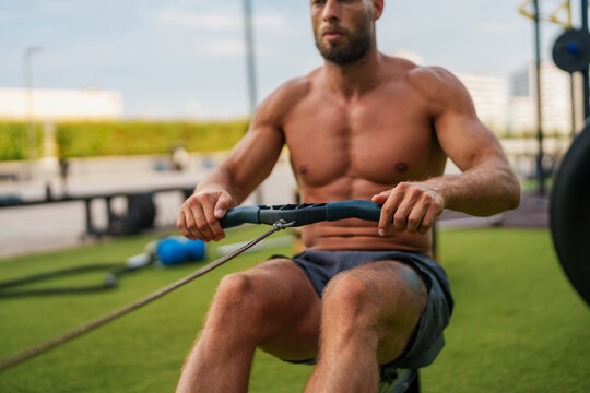 A muscular man working out on a rowing machine in an outdoor gym.