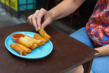 A person relishes freshly made spring rolls served with dipping sauce at a vibrant Thai street food stall.