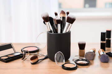 Holder with makeup brushes and different decorative cosmetics on wooden table at home