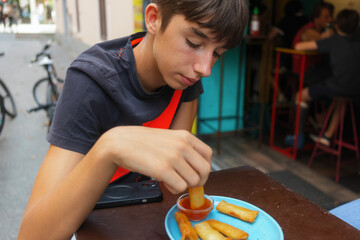 A young boy tastes crispy spring rolls with sauce at a lively Thai street food stall during a sunny...