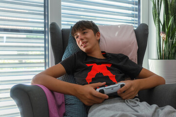 A young teen gamer boy is relaxing in a chair, playing video games and smiling in living room.