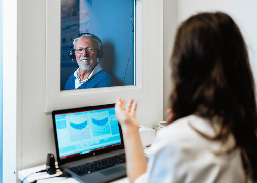 Audiologist is performing a hearing test on a smiling senior man wearing headphones inside a soundproof booth, analyzing results on a laptop