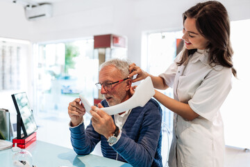 Audiologist is helping a senior man put on a hearing aid. While he looks in a mirror to see the...