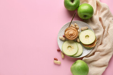 Plate with fresh apple rounds and bowl of sweet peanut butter on pink background