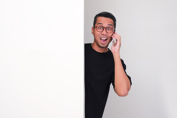 Adult asian man peeking behind wall while answering a phone call