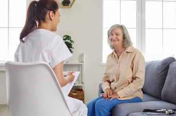 Obraz premium Friendly nurse or doctor visits senior patient at home. Young medical worker in uniform with clipboard sitting on chair and talking to happy smiling old woman on couch. Geriatrics, health care concept