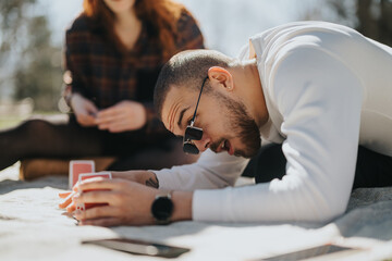 A youthful couple enjoys a game of cards together, relaxing on a blanket outside in a sunlit park setting during springtime.