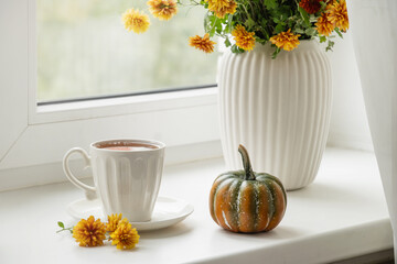 Autumn tea in white cup, decorative pumpkin and orange fall flowers on white window sill. Cozy home interior and calm. Close up.