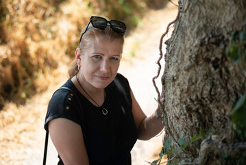 A woman standing near a tree outdoors, with natural light, showcasing relaxed and thoughtful expression. Middle-aged woman walking on footpath, wants to be in good shape