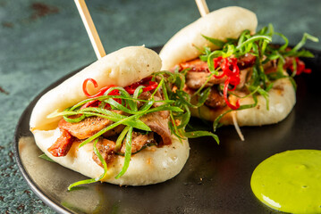 Two Asian bao buns with grilled pork, chili, and green onions on a dark plate with green sauce. Street food presentation on a textured green background