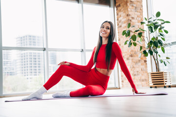 Fototapeta premium Smiling woman in red sportswear practicing yoga at home in a stylish loft space with natural light and brick walls