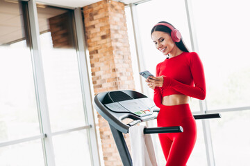 Athletic Woman in Red Fitness Outfit Using Phone While Exercising on a Treadmill in a Modern Gym...