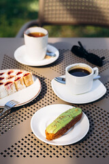 Pistachio eclair, cake slice, and coffee on a table in garden terrace setting.