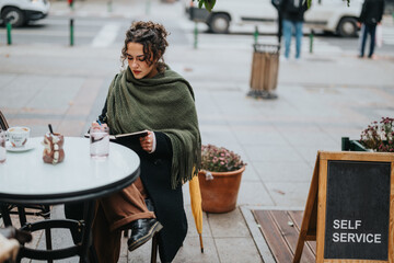 Young woman wrapped in a green shawl enjoys reading and writing while sitting outdoors at a cafe....