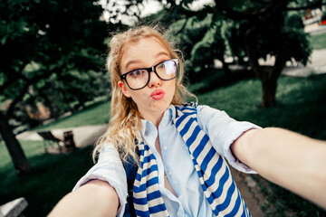 Charming girl wearing glasses and striped scarf taking a playful selfie in an outdoor urban park...