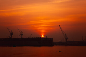 Obraz premium sunset harbor view. The bright orange and red hues reflect on the water. Silhouetted cranes and a large ship