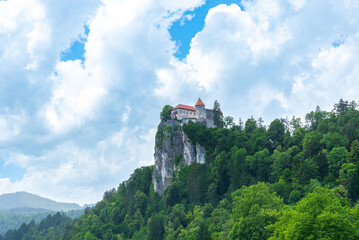 Historic Bled Castle perched on cliff above forest with cloudy summer sky in background