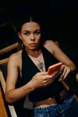 Close-up of a young woman with intense eyes, holding a phone. She is dressed in a black halter top and jeans, her green nails and jewelry adding a bold accent to the dark background.