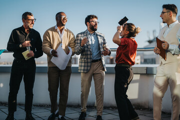 A group of multiracial business people celebrate together on a high-rise balcony at sunset, exuding...