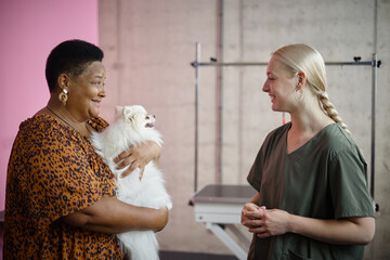 Middle aged Black woman holding fluffy dog while smiling at young adult Caucasian woman with blonde hair standing nearby in veterinary clinic setting