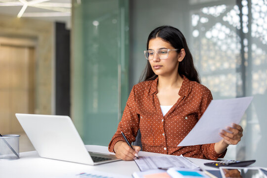 Serious and pensive business woman behind paper work inside office, female financier worker thinks about contracts and reports with charts and graphs, successful woman uses laptop at work.