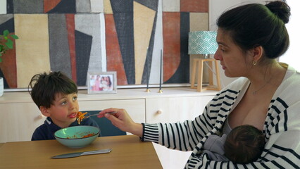 Woman feeding spaghetti to her son while breastfeeding newborn in a cozy home environment,...