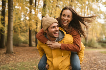 Fototapeta premium Couple laughing piggyback ride in autumn forest with yellow leaves fall